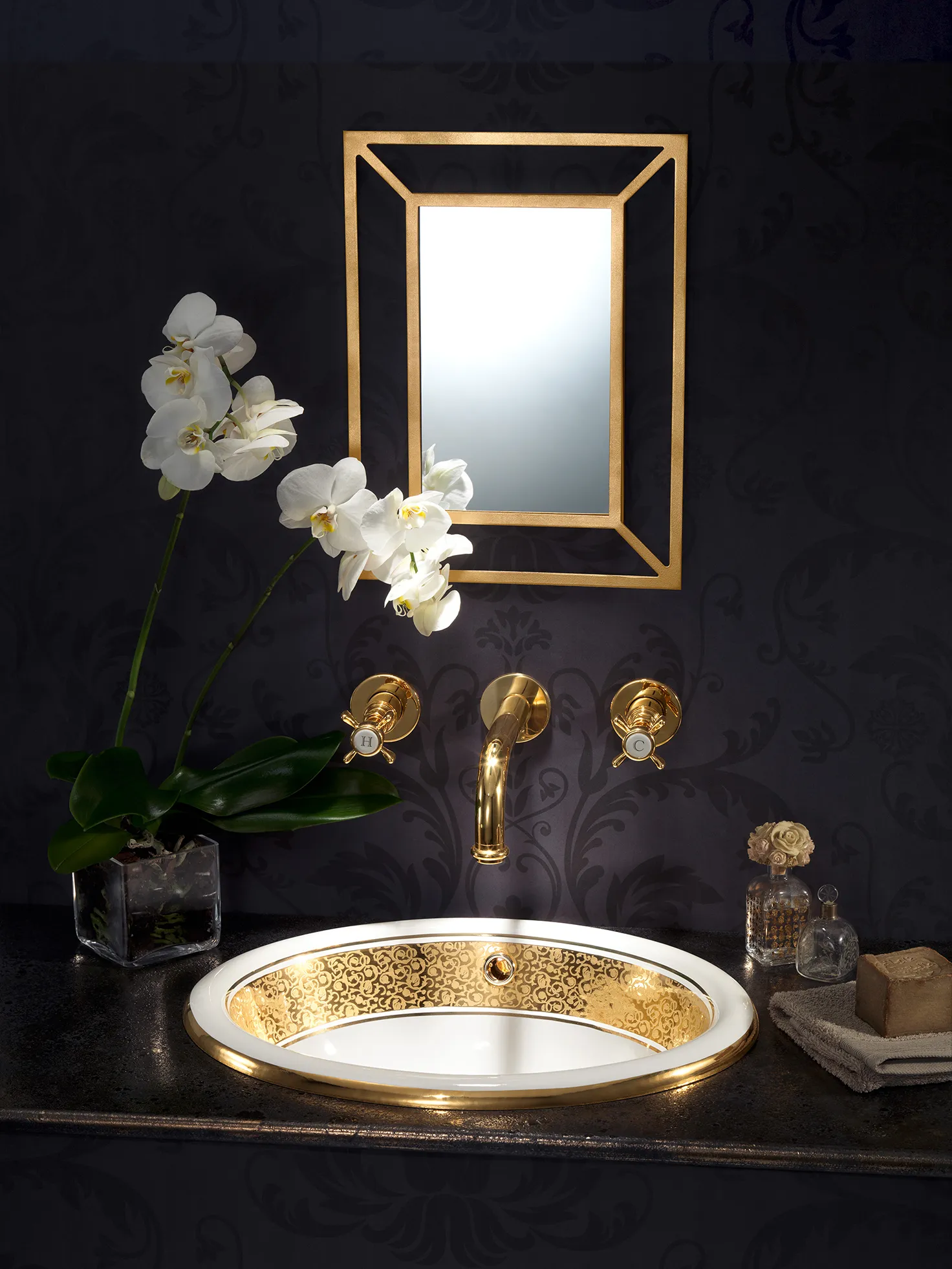 lavabo in oro e ceramica bianca in un bagno con carta da parati scura e floreale. Rubinetteria a parete oro. Specchio oro.